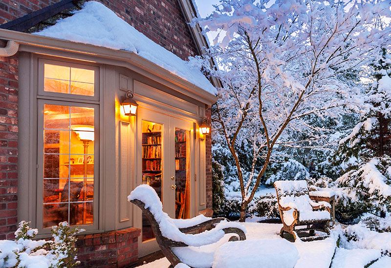 An exterior view of a snow covered home with a large patio door on a winter's evening.