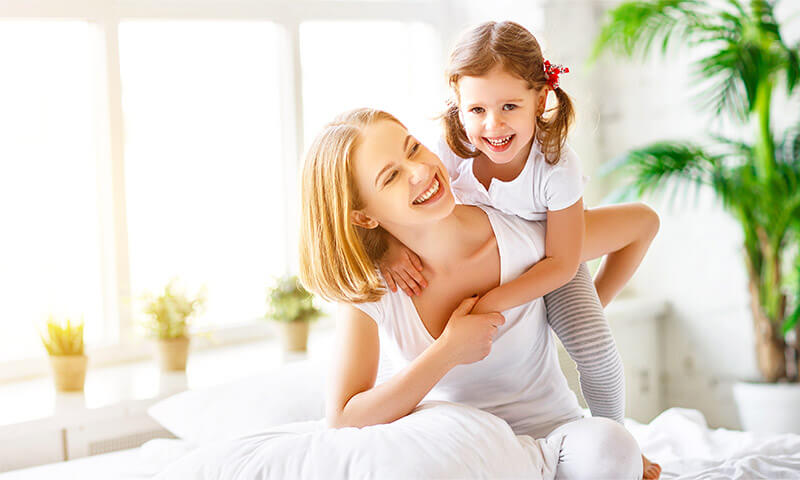 A mother playing with her daughter in their bedroom with a large bay window in the background.